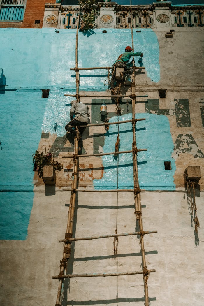 Two men painting a building using a bamboo ladder, highlighting traditional renovation techniques.